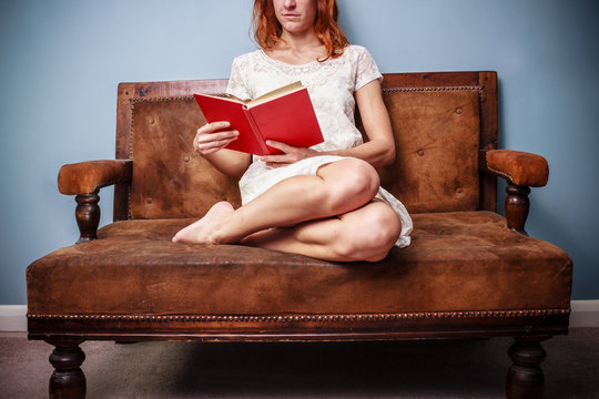 Young woman in summer dress is sitting on old sofa and reading