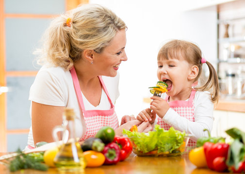 Mother Feeding Kid Daughter Vegetables In Kitchen