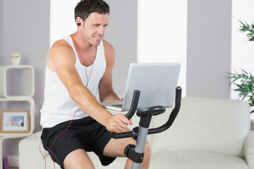 Naklejka na ściany i meble Sporty man with earphones exercising on bike and holding laptop