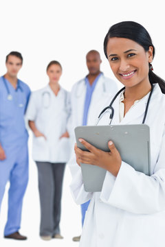 Smiling Female Doctor With Clipboard And Staff Behind Her