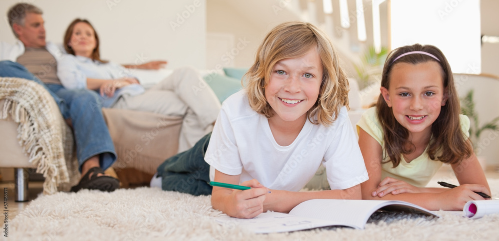 Poster siblings doing their homework on the floor with parents behind t