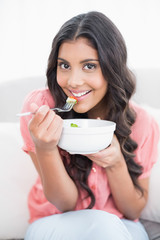 Gleeful cute brunette sitting on couch holding salad bowl