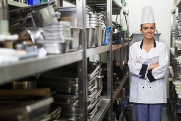 Young happy chef standing arms crossed between shelves