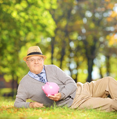 Naklejka na ściany i meble Satisfied mature gentleman in a park holding a piggy bank