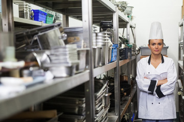 Young frowning chef standing arms crossed between shelves