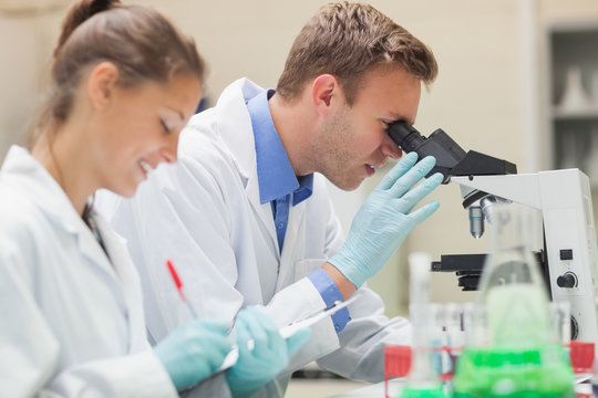 Two happy students looking through microscope and taking notes
