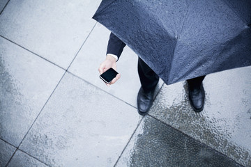 High angle view of businessman holding an umbrella and looking at his phone in the rain