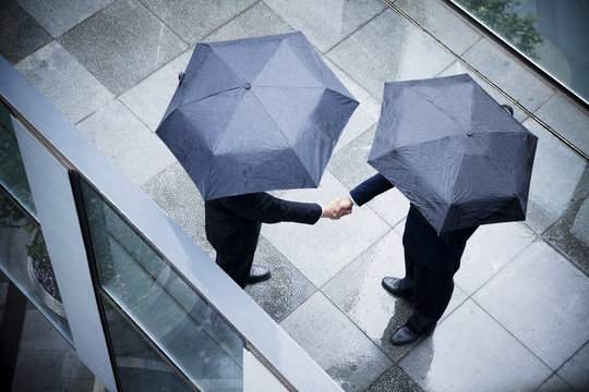High angle view of two businessmen holding umbrellas and shaking hands in the rain - Powered by Adobe