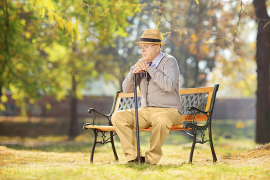 Sad Senior Man With Cane Sitting On A Wooden Bench In A Park