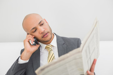 Elegant businessman using cellphone while reading newspaper
