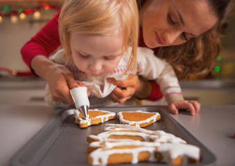 Baby helping mother decorate christmas cookies with glaze - obrazy, fototapety, plakaty