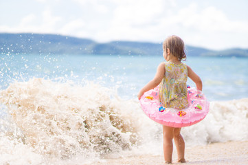 Adorable girl with inflatable ring stay in front of waves on bea