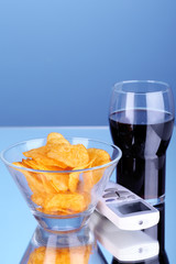 Chips in bowl, cola and TV remote on blue background