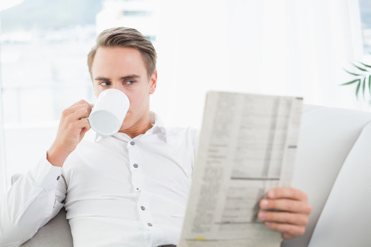 Relaxed man drinking coffee while reading newspaper on sofa - Powered by Adobe