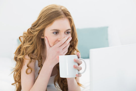 Shocked casual blond drinking coffee while using laptop in bed