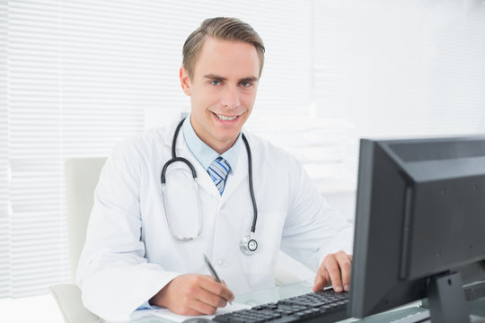 Smiling doctor writing note while using computer at medical offi