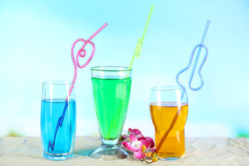 Glasses of cocktails on sand, on bright background