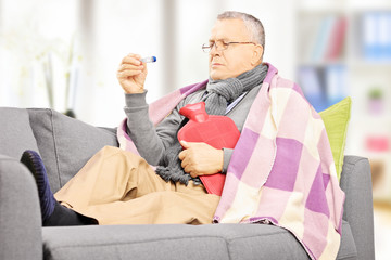 Sick man on a sofa with hot-water bottle looking at thermometer