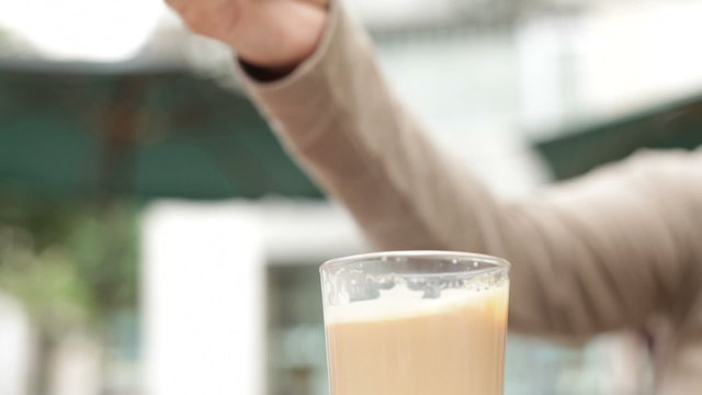 Hand with spoon stirring and pouring glass of coffee with milk.
