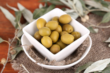 Olives in bowl with branch on sackcloth on wooden table