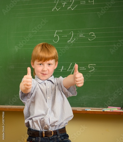Little funny redhead schoolboy near blackboard with thumbs up