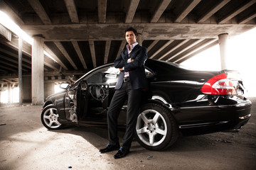 man in suit leaning against black expensive car under bridge