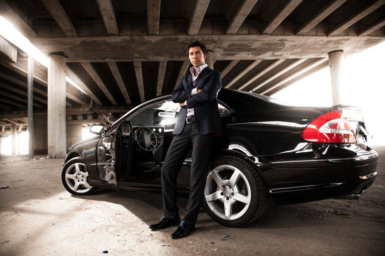 Man In Suit Leaning Against Black Expensive Car Under Bridge