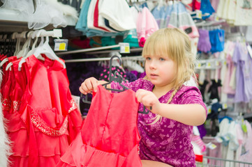 Adorable girl on shoppping cart select red dress in supermarket