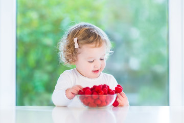 Happy smiling baby girl eating raspberry at a white table