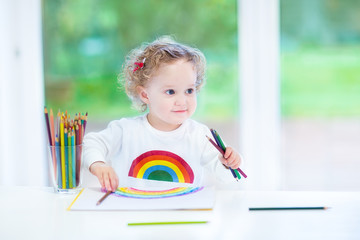 Funny smiling toddler girl drawing a rainbow at a white desk