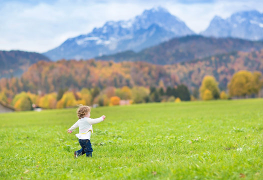 Little Toddler Girl Running In A Field Between Snow Mountains