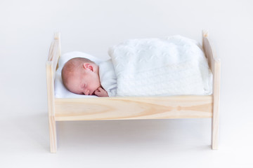 Tiny newborn baby sleeping in a toy crib under a white blanket