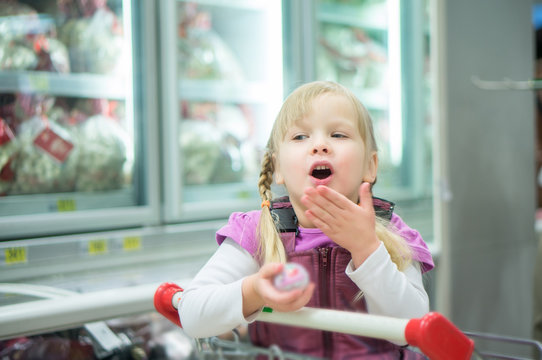 Adorable girl sit on shopping cart in front of fridges