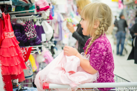 Adorable girl on shoppping cart select pink dress in supermarket