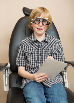Boy With Trial Frame Holding Test Chart At Optician