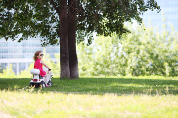 Little cute girl rides on toy car on grass near big trees