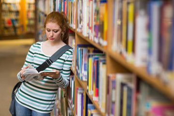 Redhead student reading book leaning on shelf in library