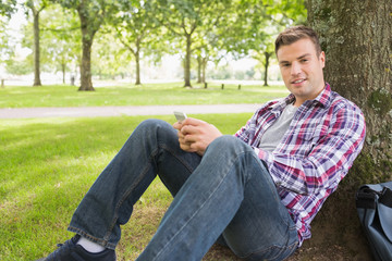 Cheerful student sending a text outside leaning on tree