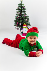 Baby boy dressed as Santa's Helper lying next to Christmas tree.