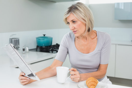 Woman Drinking Coffee While Reading Newspaper In Kitchen