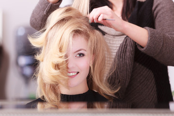 Fototapeta premium Smiling girl with blond wavy hair by hairdresser in beauty salon