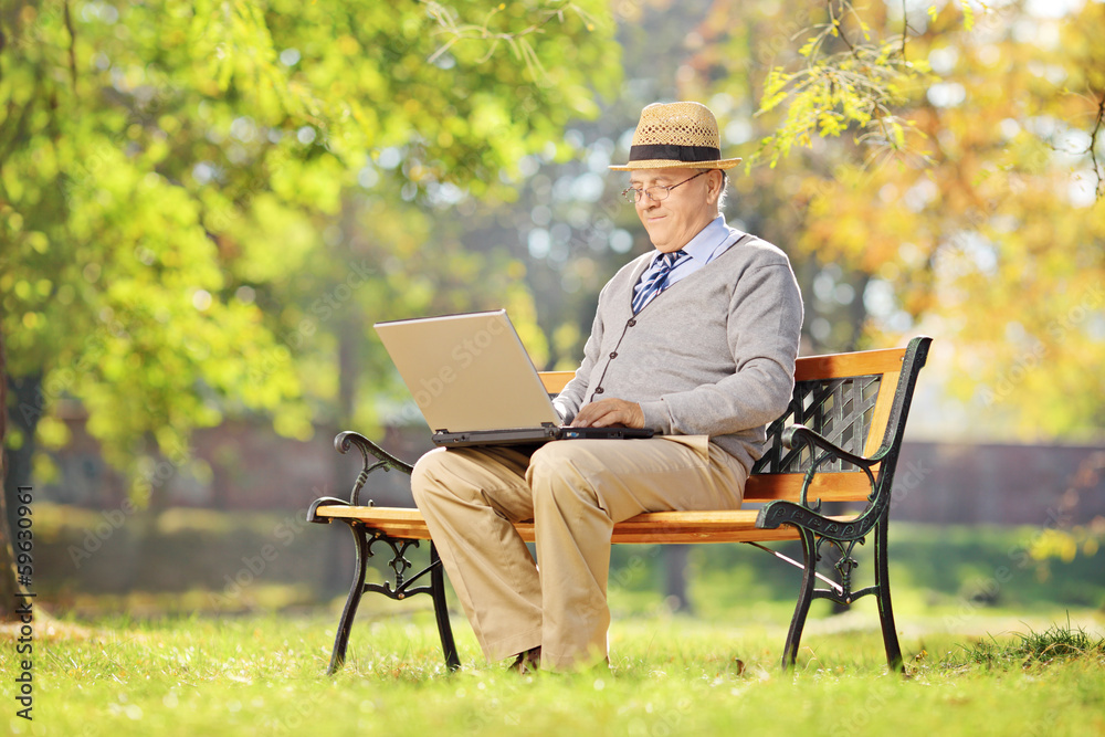 Poster Senior man with hat sitting on a bench and working on laptop