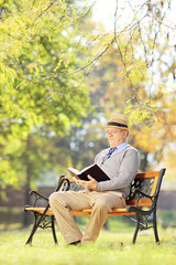 Senior man with hat seated on a wooden bench reading a book