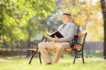 Senior man with hat sitting on a bench and reading a novel, in a
