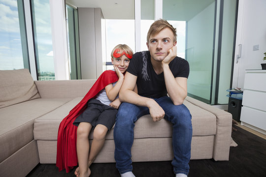 Portrait of smiling boy dressed in superhero costume sitting with sad father on sofa bed at home