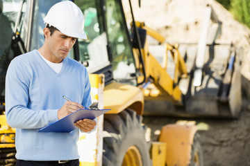 Male architect writing on clipboard against earthmover at construction site