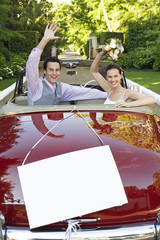 Happy newlyweds waving in convertible with blank sign in foreground