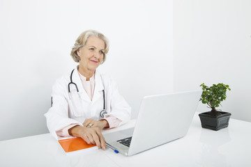 Thoughtful senior female doctor with laptop at desk in clinic