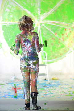 Naked girl covered in paint standing in studio with painted wall