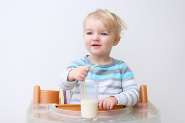 Cute blonde toddler girl drinking milk from glass with straw
